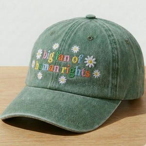 May include: Green baseball cap with the embroidered text "big fan of human rights" in colorful letters. The cap features small white daisy accents and a curved brim. A casual accessory for everyday wear.