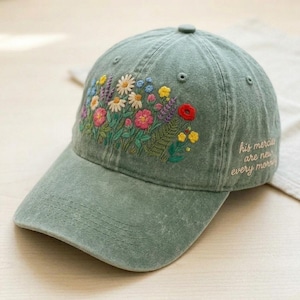 May include: A sage green baseball cap with an embroidered floral design. The flowers include pink, red, yellow, and white blooms. The text "his mercies are new every morning" is embroidered on the side.