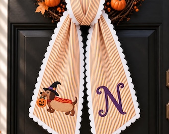 May include: A hanging door decoration featuring two long, scalloped-edge fabric panels with orange and white vertical stripes. One panel displays an embroidered dachshund wearing a witch hat and hot dog costume, while the other features a purple letter N.