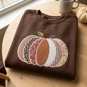 May include: A dark brown crewneck sweatshirt featuring a large, central pumpkin applique. The pumpkin is composed of vertical fabric segments in various patterns, including leopard print, floral, polka dots, and brown gingham checks.