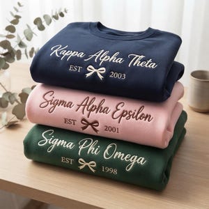 May include: A stack of three crewneck sweatshirts in navy blue, light pink, and dark green. Each features embroidered text: Kappa Alpha Theta EST 2003, Sigma Alpha Epsilon EST 2001, and Sigma Phi Omega EST 1998, all accented with a small embroidered bow.