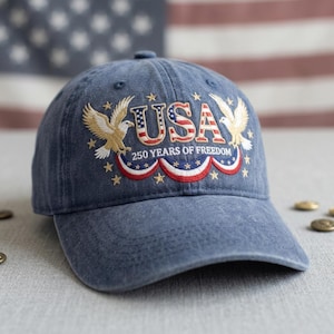 May include: A blue baseball cap with the word "USA" embroidered in red, white, and blue, flanked by golden eagles. The text "250 YEARS OF FREEDOM" is below the word "USA". The cap has a curved brim and is set against a blurred American flag.