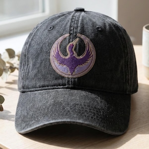 May include: A dark gray baseball cap with a curved brim. The cap features a circular embroidered patch with a purple bird design. The bird is set against a light purple background with a gold border. The cap is made of a soft, worn material.