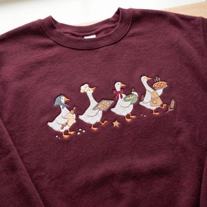 May include: A maroon crewneck sweatshirt featuring an embroidered design of four white geese engaged in baking activities. The geese are depicted wearing accessories like a headscarf, chef hat, and apron, while holding items such as a rolling pin, a tray of cookies, a mixing bowl, and a pie.