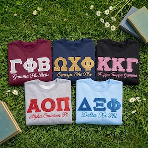 May include: Five crewneck sweatshirts in maroon, navy, black, gray, and light blue. Each features Greek letters and the corresponding organization name in glittery or textured lettering: Gamma Phi Beta, Omega Chi Phi, Kappa Kappa Gamma, Alpha Omicron Pi, and Delta Xi Phi.