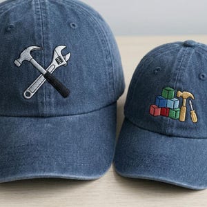 May include: Two blue denim baseball caps. The larger cap features an embroidered design of a crossed hammer and adjustable wrench in silver and black. The smaller cap features an embroidered design of colorful building blocks, a hammer, and a screwdriver.