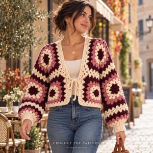 May include: A cropped cardigan made of cream, dusty rose, and dark maroon crochet granny squares. The garment features long sleeves with ribbed cuffs, an open front with a tie closure, and a V-neckline, worn over a plain white top.