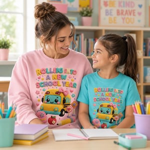 以下が含まれることがあります： 薄いピンクの長袖スウェットシャツとターコイズブルーの半袖Tシャツ。どちらもピンクのリボンをつけた黄色いスクールバス、ピンクのバックパック、リンゴ、鉛筆のグラフィックが描かれ、Rolling Into A New School Yearというテキストが添えられています。
