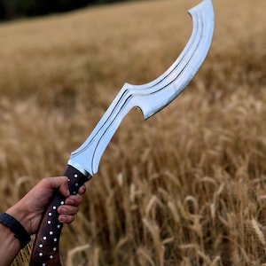 May include: A curved, silver-bladed knife with a dark wooden handle featuring white dot accents. The blade has a distinctive shape and is held against a backdrop of golden wheat fields.