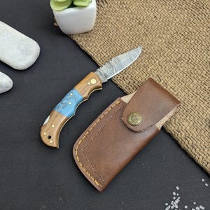 Puede incluir: Un pequeño cuchillo de bolsillo plegable con una hoja estampada y un mango de madera con detalles azules. El cuchillo está junto a una funda de cuero marrón con cierre de botón a presión. Una pequeña planta suculenta en una maceta blanca está en el fondo.