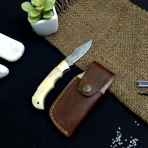 Puede incluir: Un pequeño cuchillo plegable decorativo con una hoja estampada y un mango color crema. El cuchillo está junto a una funda de cuero marrón con un cierre de botón decorativo. Una pequeña planta suculenta en una maceta blanca también está en la imagen.