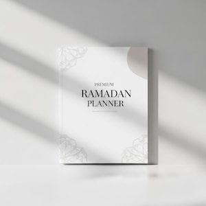 Puede incluir: Un planificador del Ramadán blanco con la palabra "PREMIUM" encima de las palabras "RAMADAN PLANNER" en negro. El planificador tiene un diseño decorativo en las esquinas y una sombra suave en el lado derecho.
