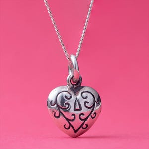 Op de afbeelding: Een zilveren hartvormige hangerketting met een sierlijk ontwerp. De hartvormige hanger hangt aan een fijne ketting. De hanger heeft een gedetailleerd patroon van krullen en een driehoekige vorm. De achtergrond is effen roze.