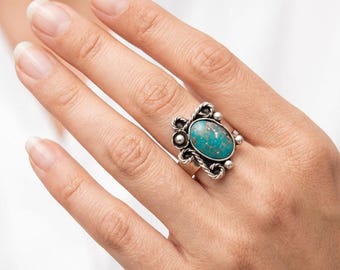 Puede incluir: Un anillo de plata con una piedra turquesa ovalada. El anillo tiene un engaste decorativo de plata con detalles de cuerda y cuentas. El anillo se muestra en una mano.