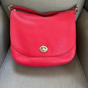 May include: A bright red shoulder bag with a textured surface and a curved flap closure. The bag features a silver-toned metal turn-lock clasp centered on the front flap and is attached to a matching red shoulder strap with silver hardware.