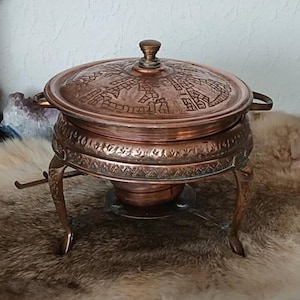 May include: A copper-colored chafing dish with a lid featuring an etched design. The dish sits on a decorative stand with curved legs. The lid has a small knob. The chafing dish is designed for serving food.