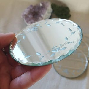 May include: A clear, round glass coaster with a scalloped edge and etched floral design. The coaster has a light blue tint and is held in a hand. Other coasters are visible in the background, along with amethyst and green crystals.