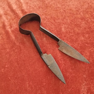 May include: Antique metal shears with a loop handle and two blades. The shears are dark brown and appear to be made of iron. The blades are pointed and slightly curved. The shears are lying on a red velvet surface.