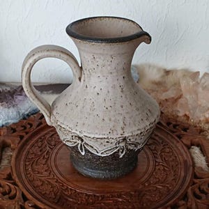 Op de afbeelding: Een gespikkelde beige keramische kan met een donkerbruine rand en handvat. De kan staat op een gesneden houten dienblad met een decoratief bloemmotief. De basis van de kan is donkerbruin met een witte decoratieve band.