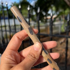 May include: A tan ceramic wand with dark blue, oblong spots. The wand is cylindrical and held in a hand. The background includes a black metal fence, a white wall, and a blue sky.