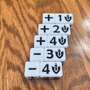 May include: White game dice with black numbers and symbols, including plus and minus signs, and a sword and shield icon. The dice are stacked on a wooden surface.
