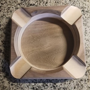 Könnte beinhalten: Quadratischer Aschenbecher aus Holz mit kreisförmigem Innenraum und vier Eckablagen. Der Aschenbecher besteht aus hellem und dunklem Holz mit natürlicher Oberfläche. Der Aschenbecher ist für Zigaretten oder Zigarren konzipiert.