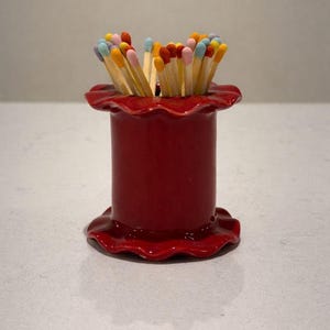 Puede incluir: Un portafósforos de cerámica roja con la parte superior e inferior con volantes, lleno de fósforos. Los fósforos tienen puntas de colores en tonos rosa, naranja, amarillo y azul. El soporte es cilíndrico y está sobre una superficie clara.