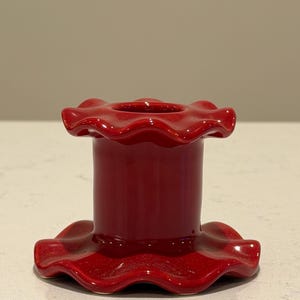 Può includere: Un portacandele in ceramica rossa lucida con un bordo increspato e smerlato. Il portacandele ha un corpo cilindrico e una base ampia, anch'essa con un bordo increspato. Il design è semplice ed elegante, adatto a vari stili di arredamento.