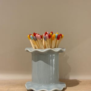 May include: A pale blue ceramic match holder with a frilled edge, filled with matches. The match heads are various colours, including red, yellow, pink, and blue. The holder is on a wooden surface.