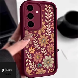 Puede incluir: Funda de teléfono burdeos con un estampado floral de flores naranjas, amarillas y blancas y hojas verdes. La funda tiene un módulo de cámara con tres lentes. La marca "CASETEC" es visible.