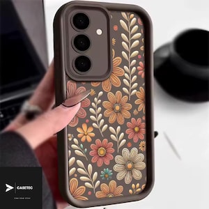 Puede incluir: Una funda de teléfono marrón con un estampado floral de flores naranjas, rosas y amarillas, y hojas blancas. La funda tiene un borde marrón oscuro y un módulo de cámara. La marca "CASETEC" es visible.