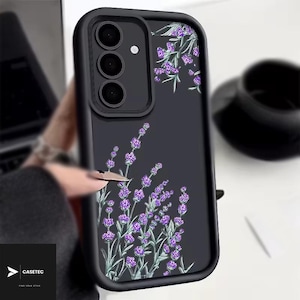 Può includere: Custodia per telefono nera con un design floreale di lavanda. La custodia ha un bordo nero e un retro trasparente con un'illustrazione di lavanda viola e verde. La custodia ha un modulo fotocamera con tre obiettivi.