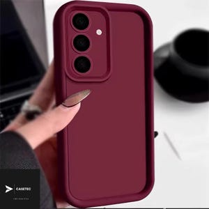 May include: A solid deep burgundy smartphone case featuring a raised rectangular camera module with three circular lens cutouts and a small circular flash opening. The case has a smooth matte finish and a thick, protective border frame.