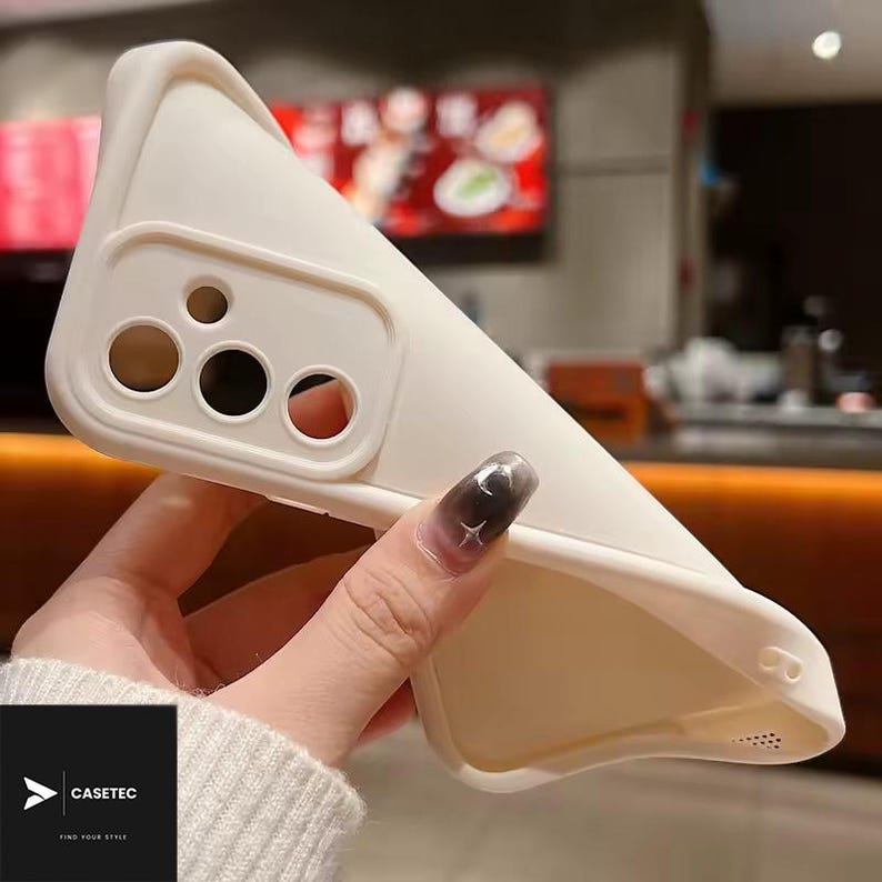 May include: A cream-colored phone case with cutouts for the camera and other features. The case is held in a hand, showcasing its flexible design. The brand name "CASETEC" is visible in the lower left corner.