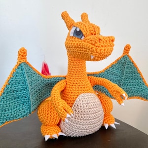 May include: Crocheted orange dragon with teal wings and a beige belly. The handmade amigurumi figure has white claws and blue eyes. A unique, handcrafted plush toy.