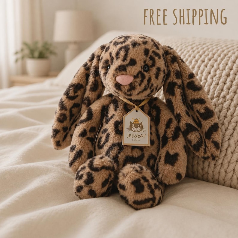 May include: A plush stuffed animal in the shape of a rabbit with long ears, covered in a brown and black leopard print pattern. The rabbit has a soft pink nose and a small tag around its neck that reads Jellycat London with a cat icon.