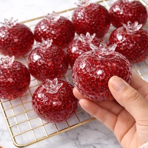 May include: A collection of decorative glass pomegranate ornaments. Each pomegranate is a deep red color with a textured surface and a clear, crown-like top. The pomegranates are displayed on a gold wire rack.