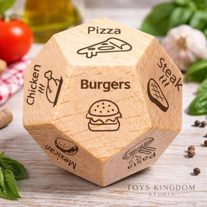 May include: A wooden, multi-sided die featuring food illustrations and labels. Sides include pizza, burgers, steak, pasta, Mexican, and chicken. The die is light brown with dark brown graphics and text. The Toys Kingdom Studio logo is at the bottom.