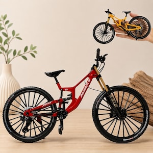 May include: A miniature red mountain bike model with black wheels, a black seat, and gold-toned front suspension forks. The frame features white text detailing. A second, smaller yellow mountain bike model is shown held in the upper right corner.