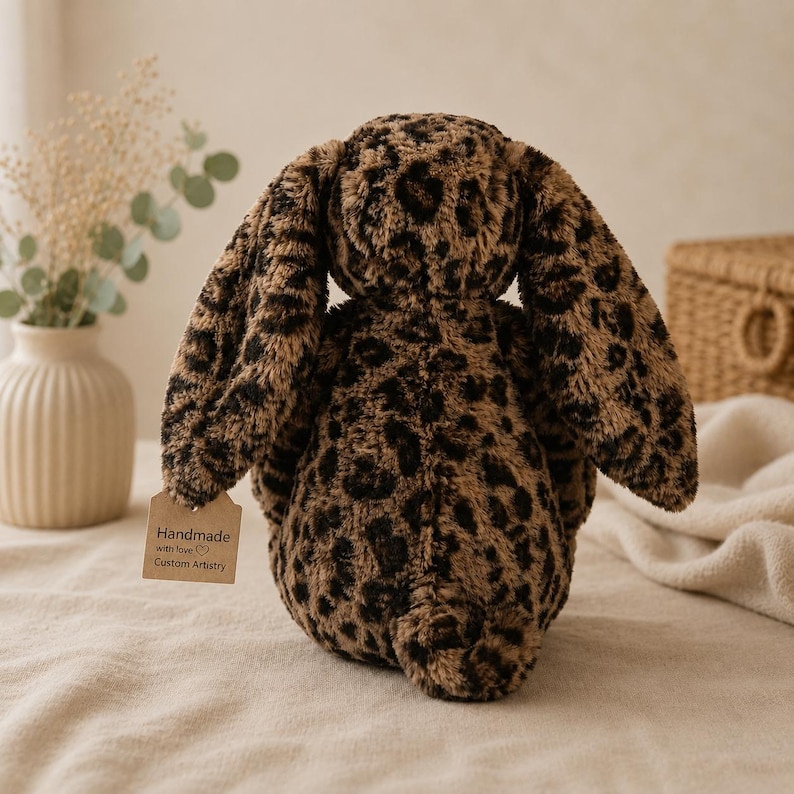 May include: A plush stuffed animal in the shape of a rabbit with long ears, covered in a brown and black leopard print pattern. A small, rectangular, light brown paper tag is attached, featuring the text Handmade with love Custom Artistry.