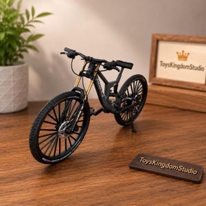 May include: A miniature black mountain bike model featuring gold-colored front suspension forks, black spoked wheels with disc brakes, and a frame marked with the text "MODERN CYCLE". The bike is displayed on a wooden surface near a small sign.