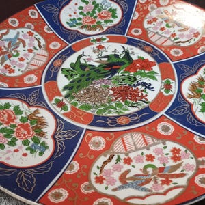 May include: A decorative ceramic plate with a vibrant design. The plate features a central image of two peacocks surrounded by floral patterns in red, blue, and white. The plate's rim has a gold trim.