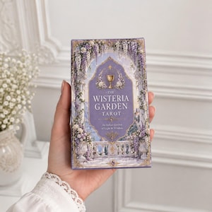Pode incluir: Uma caixa retangular de baralho de tarot intitulada The Wisteria Garden Tarot, An Italian Garden of Light & Wisdom. A capa apresenta um painel central roxo com um ícone de cálice dourado, emoldurado por arcos de pedra e flores de glicínia.