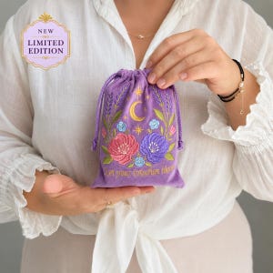 Op de afbeelding: Een paars trekkoordzakje met geborduurde roze en paarse bloemen, groene bladeren, een gele maansikkel en sterren. De tekst "Let your intuition bloom" is onderaan in gouddraad geborduurd. Een label vermeldt "NEW LIMITED EDITION".
