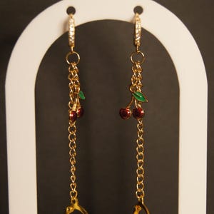 May include: Gold-toned dangle earrings featuring a chain design. Each earring has a small cluster of red cherries with green leaves. The earrings are displayed on a white arch-shaped stand against a dark background.