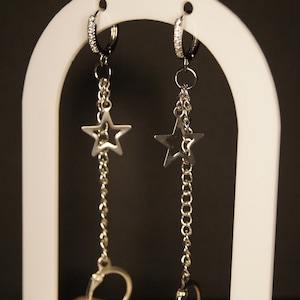 May include: A pair of silver earrings with a star and handcuff design. Each earring features a small hoop with clear stones, a chain, a star pendant, and a handcuff charm. The earrings are displayed on a white stand against a dark background.