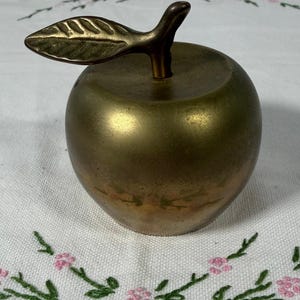 May include: A brass apple-shaped decorative object with a smooth, metallic finish and a prominent stem featuring a single leaf. The body of the apple displays a subtle, faded floral pattern in muted pink and green tones across the lower section.