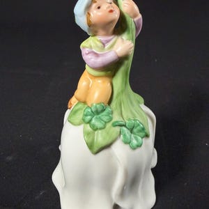 May include: A porcelain bell figurine featuring a child wearing a light blue cap, a lavender shirt, and yellow pants. The child is climbing a tall green stalk adorned with green clover leaves, which sits atop a large, white, bell-shaped flower base.