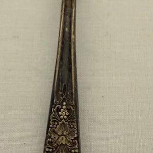 May include: A tarnished silver-toned metal spoon featuring an ornate, embossed floral and grape cluster pattern on the handle. The bowl of the spoon shows significant signs of oxidation and wear, with a dark, mottled patina covering the surface.