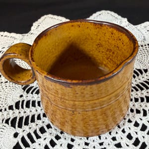 May include: A ceramic mug featuring a speckled mustard yellow glaze with a dark brown rim and a circular handle. The exterior surface has a subtle horizontal ribbed texture, and the interior is finished in the same speckled yellow tone.